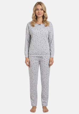 Vrouw staat blootsvoets en draagt een bijpassende lichtgrijze pyjama met een subtiel hartjespatroon en een klein zwart strikje bij de hals.
