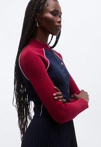 Soonikkoes kudumistega kampsun luku ja kontrastsete burgundi ning mereväe siniste paneelidega, tekstuurse kangaga ja valge aktsendiga torudel õmbluste ääres.