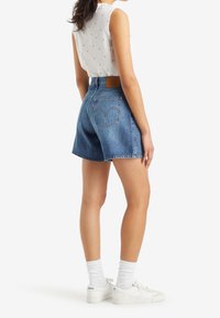 Džínové šortky středně modré barvy s vysokým pasem, zakončené třepenými lemy a hnědou koženou zadní náplastí. Kombinovány s bílými teniskami a ponožkami.