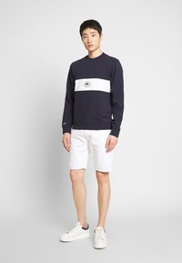 Felpa blu navy con striscia orizzontale bianca e dettagli verdi, dotata di una toppa con logo; abbinata a pantaloni corti bianchi e sneakers bianche.