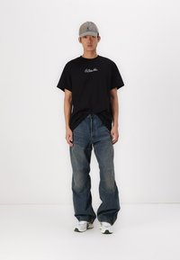 T-shirt en coton noir avec logo blanc, associé à un jean en denim bleu ample avec des détails en patchwork. Une casquette grise et des baskets blanches complètent le look.