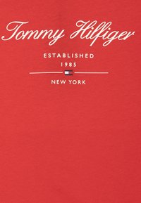 T-shirt rossa in cotone con la scritta "Tommy Hilfiger" in bianco, con "ESTABLISHED 1985" e "NEW YORK" stampati sotto, e un piccolo logo come accento.