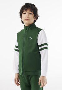 Zeleni zip-up sako s bijelim rukavima, s zelenim prugama na rukama i malim logom na prsima. Izrađen od glatkog materijala.