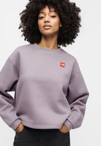 Ανοιχτό μωβ φούτερ με στρογγυλή λαιμόκοψη, ριμπ μανσέτες και μικρό κόκκινο λογότυπο στο στήθος. Υφασμάτινη υφή απαλή.