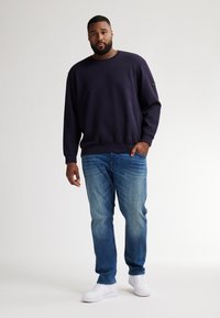 Granatowy sweter z okrągłym dekoltem i ściągaczami na rękawach, w połączeniu z jasnoniebieskimi dżinsami i białymi sneakersami, na jednolitym tle.