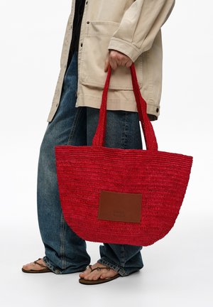 Persona sosteniendo un gran bolso tejido rojo con parche marrón, vistiendo chaqueta beige, vaqueros azules y sandalias marrones sobre fondo blanco.