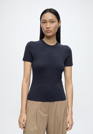 Femme aux longs cheveux noirs portant un top en maille bleu marine ajusté à manches courtes et un pantalon plissé beige taille haute, debout devant un fond blanc.