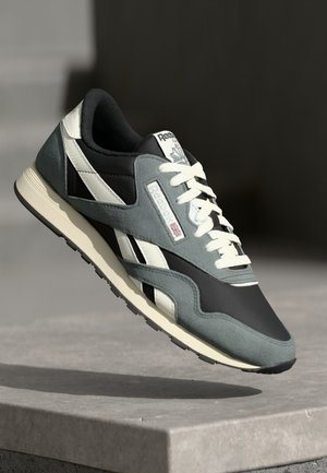 Grå og sort Reebok-sneaker med hvide snørebånd og striber, svævende over en betonoverflade mod en mørk baggrund.