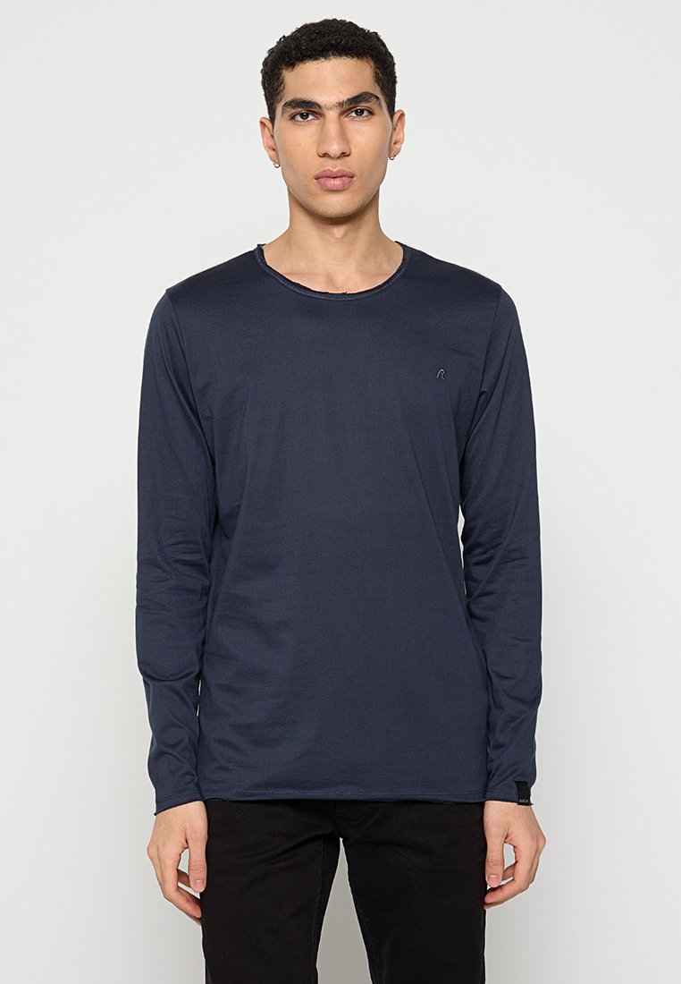 Junger Mann mit kurzen lockigen Haaren, der ein schlichtes navyblaues Langarmshirt und schwarze Hosen vor einem weißen Hintergrund trägt.