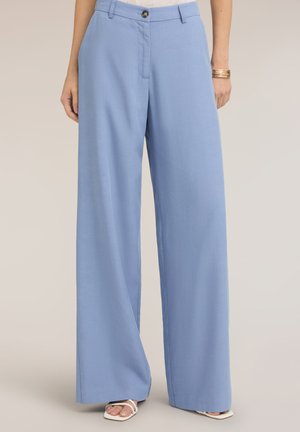 Vrouw draagt lichtblauwe wijdvallende broek, witte sandalen met vierkante neus en een gouden armband aan de rechterpols tegen een effen achtergrond.