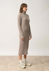 Abito in maglia color beige chiaro con scollatura alta e texture a coste. Presenta maniche lunghe e una silhouette aderente, abbinato a scarpe atletiche.