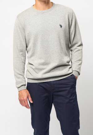 Homme portant un pull à col rond gris clair avec un petit logo sombre sur la poitrine et un pantalon bleu marine, debout avec une main dans la poche.