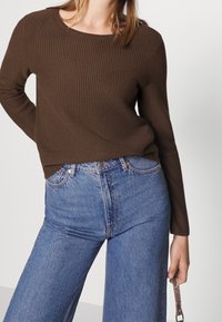 Femme portant un pull côtelé marron et un jean taille haute bleu, tenant une petite sangle à motif serpent dans sa main droite.