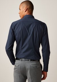 Navyblå skjorte med klassisk krave, lange ærmer og manchetknapper, kombineret med grå bukser med diskrete lommer og tekstur.