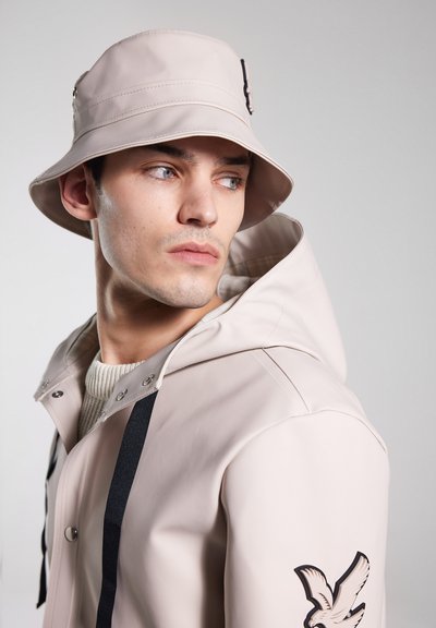 Svetlo béžový vedro klobúk s širokým okrajom, ktorý má pásik a detail s prackou. Model má na sebe zodpovedajúci pršiplášť s čiernymi akcentmi.
