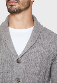 Cardigan grigio a maglia con colletto a scialle, caratterizzato da motivi a treccia, due bottoni scuri, indossato sopra una maglietta bianca.