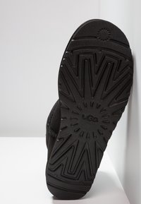 Suela negra de bota UGG con un patrón texturizado en zigzag, con acentos circulares y el logotipo de la marca grabado en el centro.