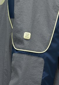 Γκρι και ναυτικά υφασμάτινα πάνελ με κίτρινη λεπτομέρεια σωληνοειδούς ραφής και ένα μικρό τετράγωνο λογότυπο στην γκρι πτέρυγα ενός ενδύματος ή τσάντας.