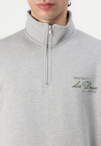 Polaire grise à col montant avec une fermeture à demi-fermeture éclair. Présente un imprimé graphique vert sur la poitrine à gauche. Texture douce.