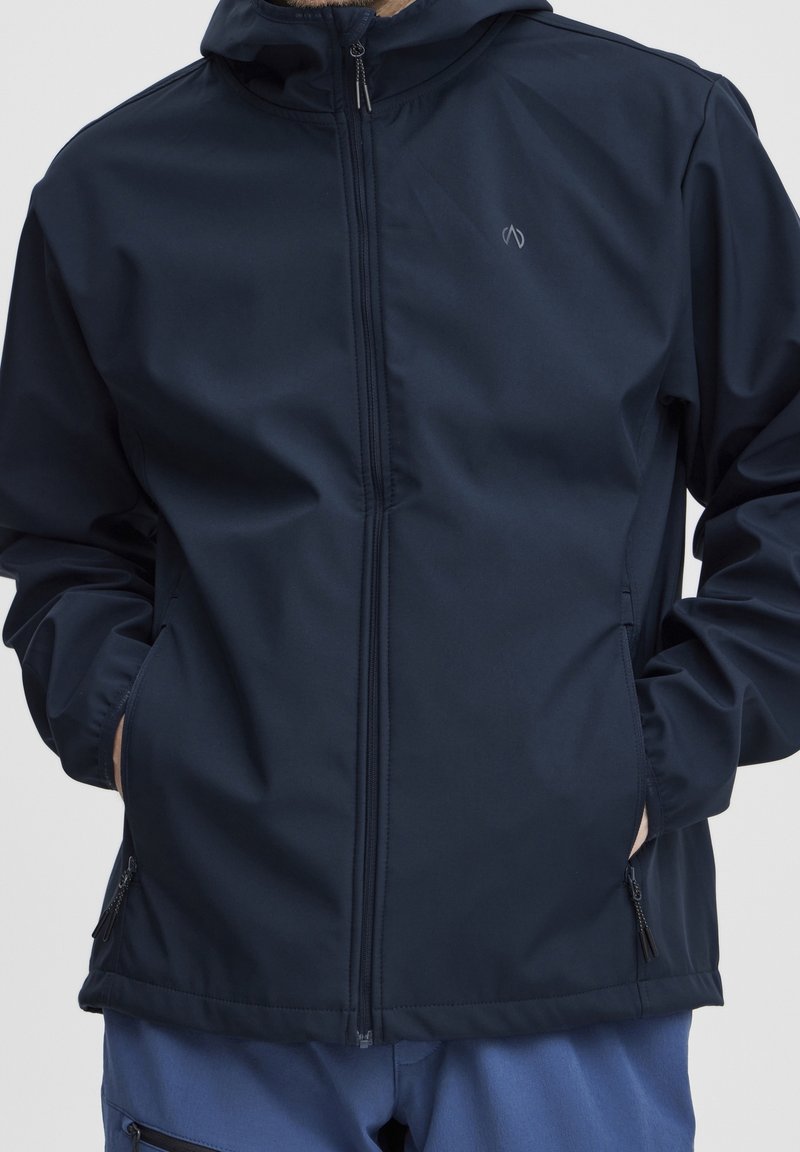 Homme portant une veste à capuche bleu marine zippée, mains dans les poches, associée à un pantalon bleu sur fond uni.