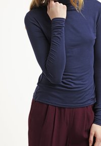 Námořnická dlouhá roláková halenka vyrobená z elastického materiálu, s úzkým střihem a nařasením v pase, v kombinaci s burgundskými kalhotami.