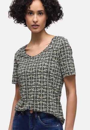 Kvinde med krøllet hår iført en sort og hvid blomstermønstret kortærmet bluse og blå jeans mod en ensfarvet baggrund.