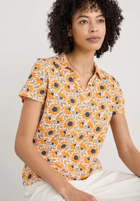 Poloko s krátkym rukávom v krémovej farbe s oranžovými slnečnicami a modrými akcentmi. Mäkká bavlna s voľným strihom a detailom v oblasti goliera.