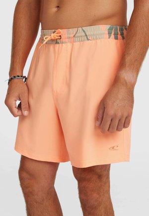 Muž v svetlo oranžových plavkách s vzorovaným pásom a šnúrkou, stojaci s rukami uvoľnene položenými popri tele.