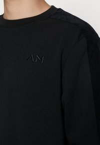 Sweat-shirt noir avec panneaux texturés côtelés aux épaules et petit logo brodé "AM" sur la poitrine, porté par une personne.