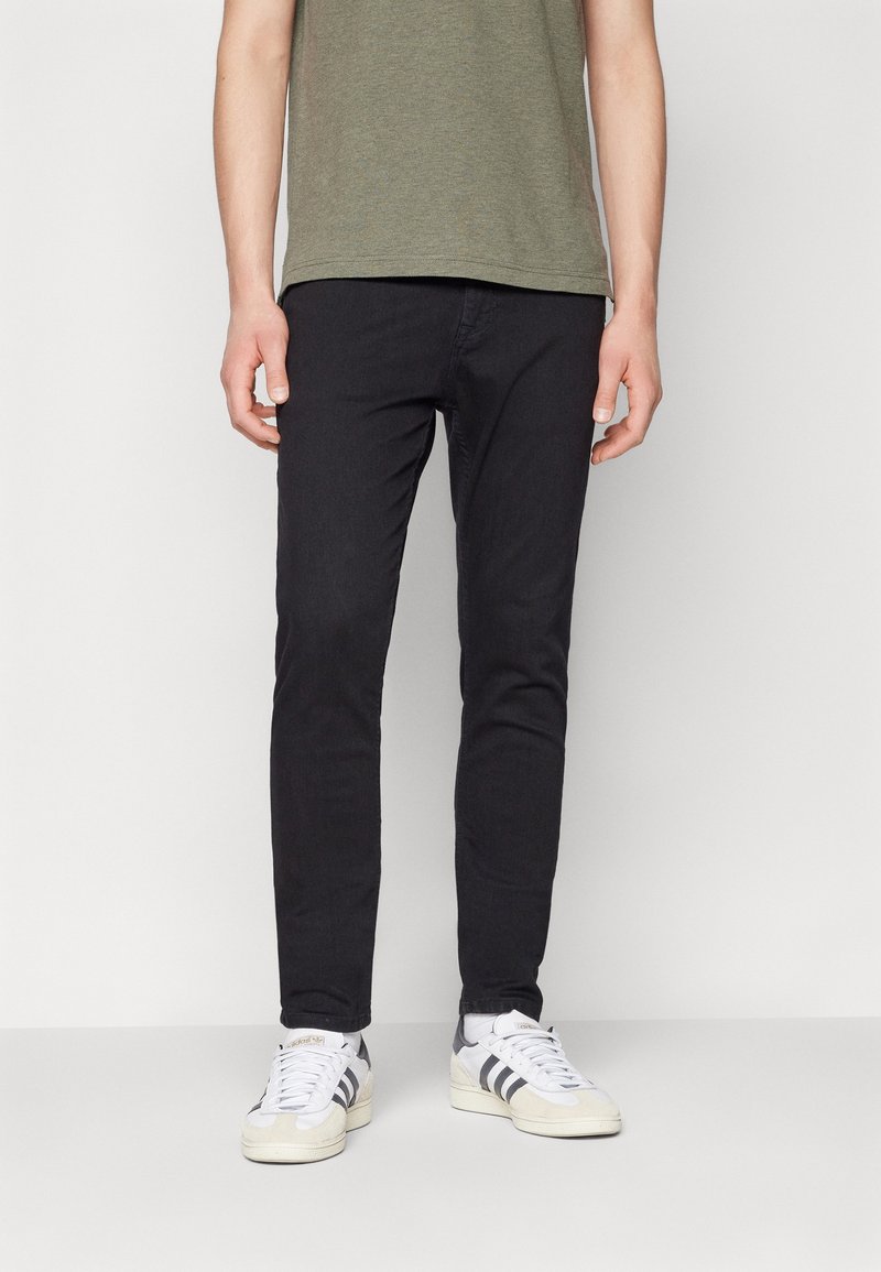 Homme portant un pantalon noir slim, des baskets blanches avec des rayures noires et une chemise à manches courtes vert olive, debout devant un fond blanc.