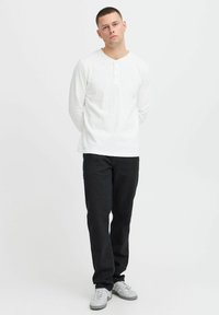 Weißes Langarm-Henley-Shirt mit drei Knöpfen, kombiniert mit schwarzen Jeans und grauen Sneakers, in entspannter Passform.