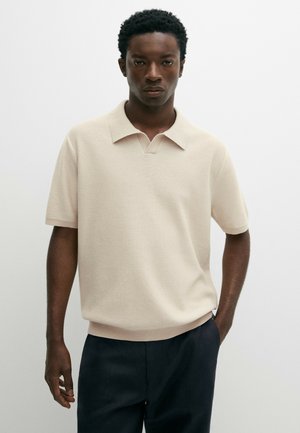 Giovane uomo con capelli corti e ricci che indossa una polo in maglia beige a maniche corte e pantaloni scuri su sfondo bianco semplice.