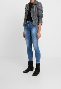 Person iført tætsiddende blå jeans, sort skjorte, grå læderjakke med lynlås åbnet og sorte ankelstøvler står op ad en ensfarvet hvid baggrund.