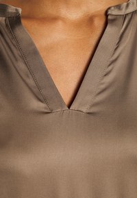 Gros plan d'une personne portant un haut taupe à col en V montrant l'encolure et la peau du haut de la poitrine.
