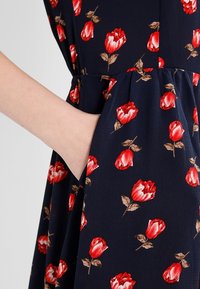 Laivastonsininen mekko, jossa on kukkakuosi punaisista ruusuista ja vihreistä lehdistä, valmistettu kevyestä kankaasta, sisältää sivutaskut.