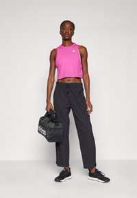 Rosa kurz geschnittenes Sporttanktop, schwarze lockere Jogginghose und schwarze Sneaker. Hält eine schwarze Sporttasche mit weißem Logo in der Hand. Einfaches Design.