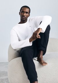 Λευκό μακρυμάνικο T-shirt με μικρό λογότυπο, συνδυασμένο με μαύρο παντελόνι φόρμας. Το μοντέλο κάθεται σε μια καρέκλα με υφή σε κρεμ απόχρωση.