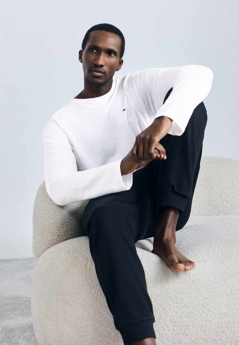 Λευκό μακρυμάνικο T-shirt με μικρό λογότυπο, συνδυασμένο με μαύρο παντελόνι φόρμας. Το μοντέλο κάθεται σε μια καρέκλα με υφή σε κρεμ απόχρωση.