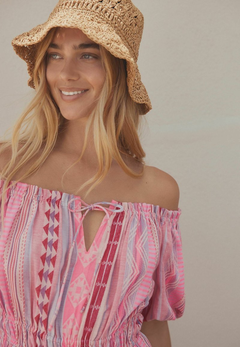 Jeune femme souriante, portant un chapeau de soleil tissé beige et une robe rose à motifs à épaules dénudées sur un fond clair uni.