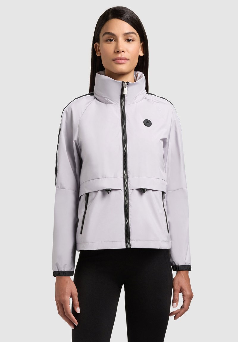 Femme portant une veste zippée gris clair avec des bordures noires et un logo sur la poitrine, associée à un pantalon noir, debout devant un fond uni.