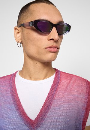 Persona con capelli corti indossa occhiali da sole cat-eye con lente violacea, orecchino a cerchio argentato, camicia bianca e gilet lavorato a maglia con sfumatura rosa-viola.