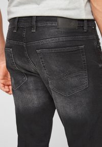 Jeans de mezclilla negra con un lavado desgastado, que cuentan con bolsillos traseros distintivos y un parche de cuero negro en la cinturilla.