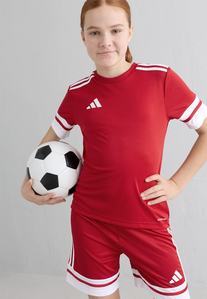 Rød atletisk skjorte og shorts med hvite detaljer, med tre hvite striper på ermene, og holder en svart og hvit fotball.