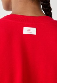 Felpa rossa con colletto a coste, dotata di un'etichetta bianca sul retro con il testo "Cool Cozy Comfy." Texture liscia.