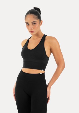 Vrouw met haar in een knot, gekleed in een zwarte mouwloze crop top en hooggetailleerde legging, staand met handen op de heupen.