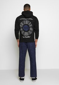 Muž v černé mikině s kapucí, na které jsou mezinárodní názvy měst a modrý globus, v kombinaci s tmavě modrými kalhotami s leopardím vzorem, otočený zády.