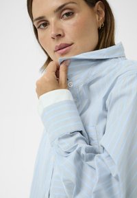 Jasnoniebieska koszula na guziki z cienkimi białymi paskami, z kołnierzykiem typu spread i białymi mankietami. Gładka faktura, casualowy design.