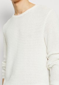 Pull en maille blanche avec un col rond, un tissage texturé et des manches retroussées, offrant un design décontracté et respirant.