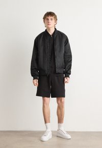 Bomber nera con finitura liscia, maglietta a girocollo e pantaloni corti neri. Sneakers bianche e calzini alla caviglia completano l'outfit.