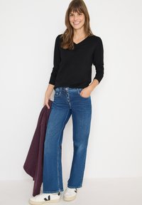 Maglietta nera a maniche lunghe, jeans blu a gamba larga, sneakers bianche con dettagli neri e un cardigan a maglia bordeaux appoggiato su un braccio.